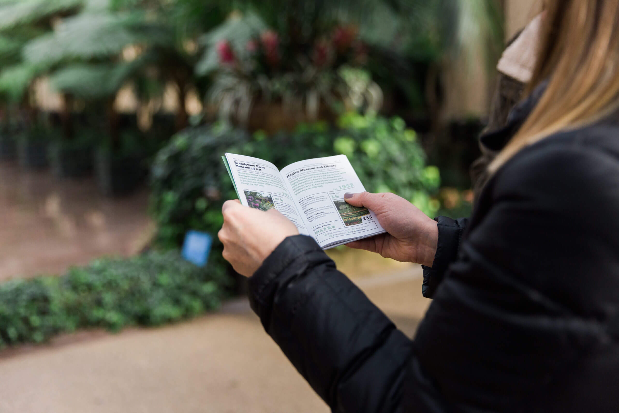 A woman at a garden wearing a black coat holding a plant description pamphlet