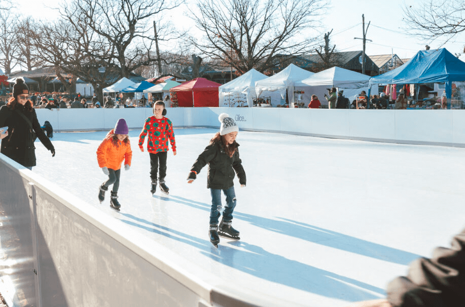 Children ice skating outdoors at a rink with tents and people in the background.