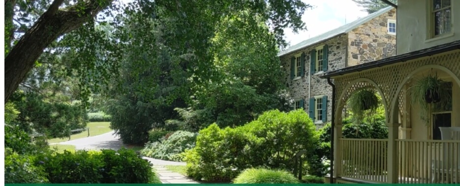 Historic stone house with green shutters at Tyler Arboretum, surrounded by lush summer trees and gardens.